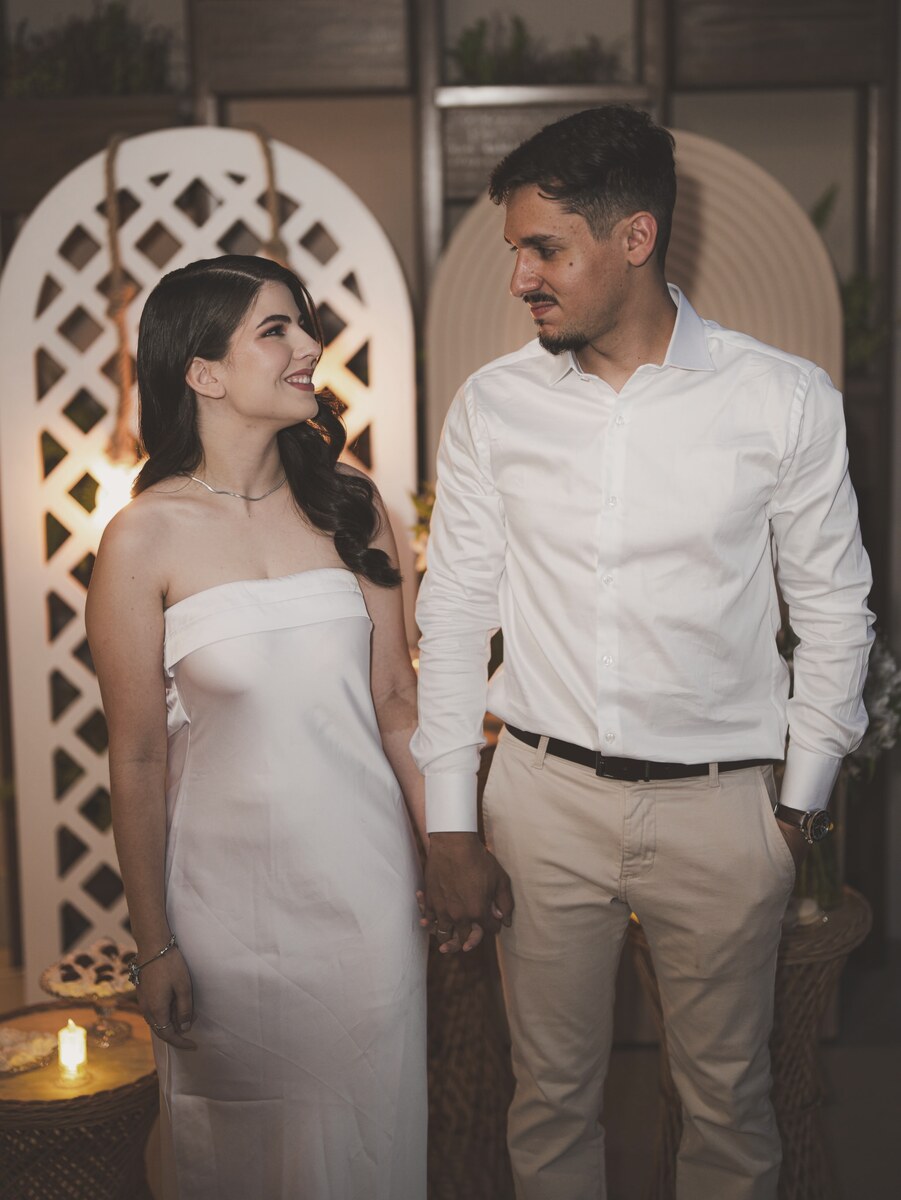 João e Mel de mãos dadas diante de um arco decorado com velas, ela de vestido claro e ele de camisa branca.