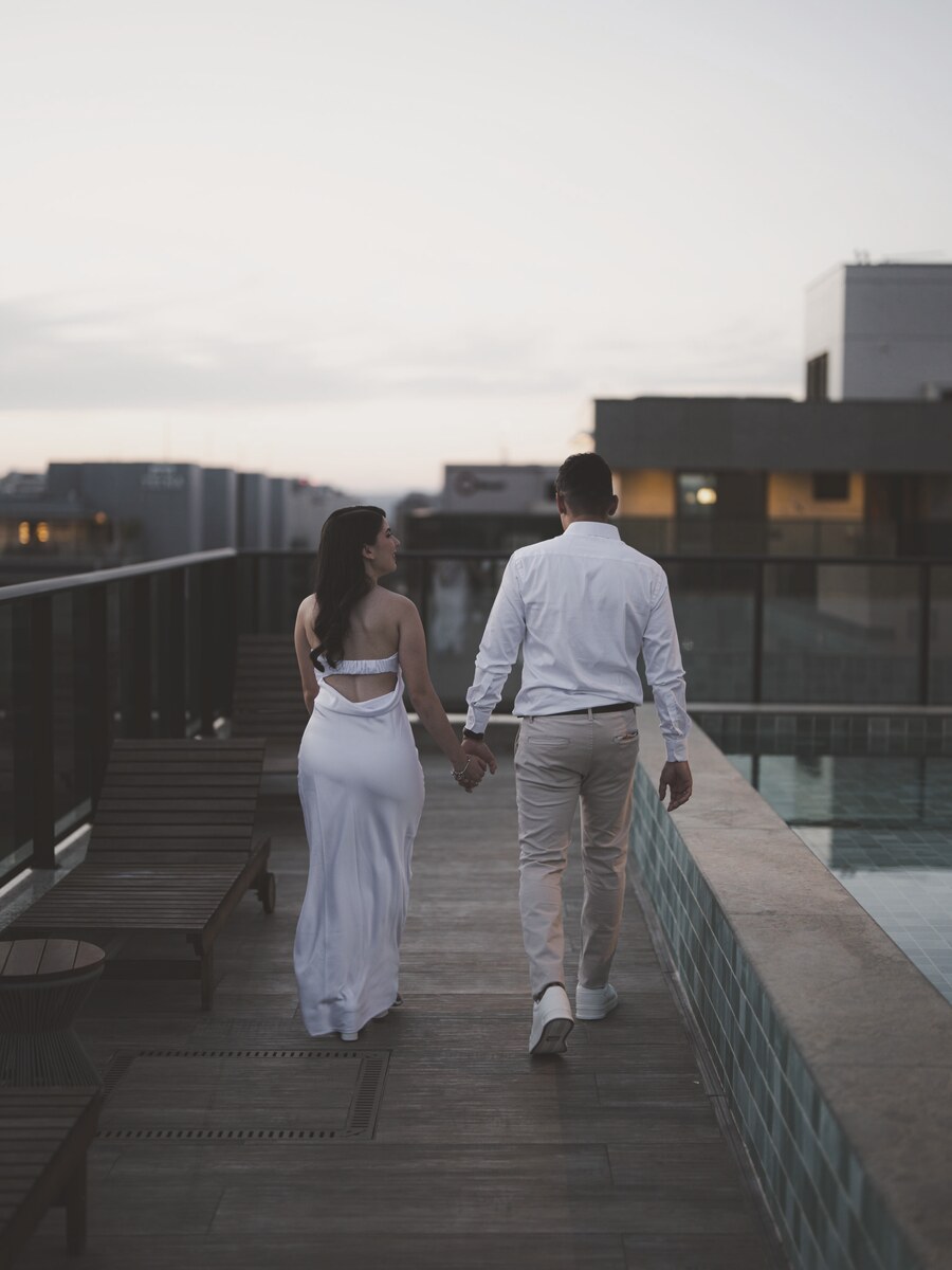 João e Mel de mãos dadas, caminhando de costas pelo deck do terraço ao pôr do sol, ao lado de uma piscina.
